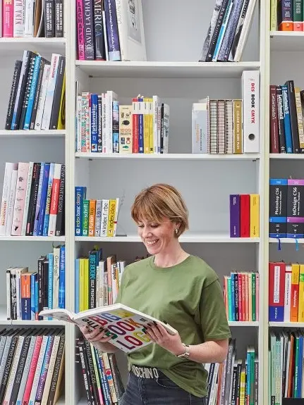 Lächelnde Frau liest in einem Buch vor einem gut gefüllten Bücherregal.“
