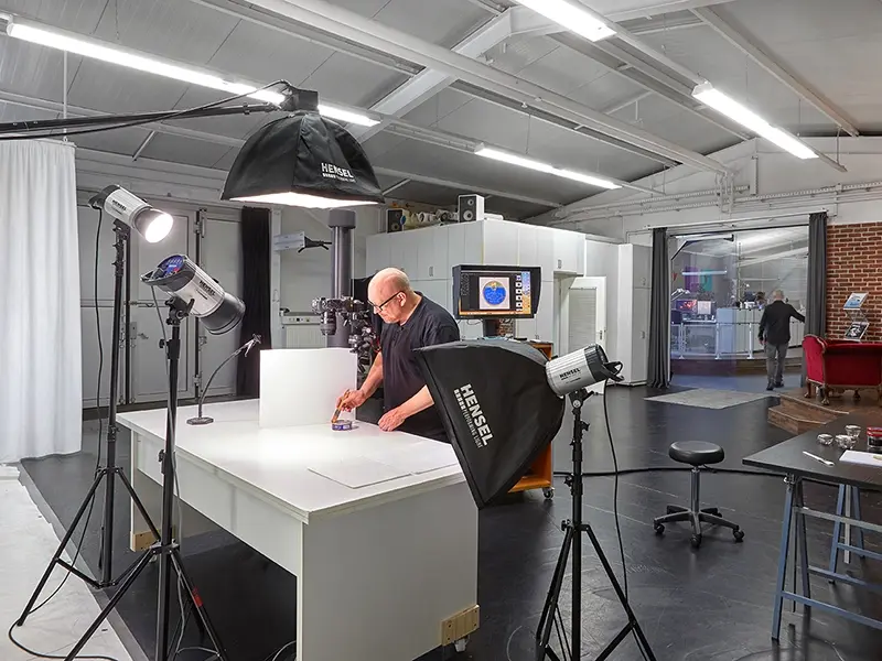 Das Bild zeigt den Fotografen Christoph Ernst in einem professionellen Fotostudio bereitet eine Produktaufnahme auf einem weißen Tisch vor, umgeben von Hensel-Studioblitzen und Softboxen.